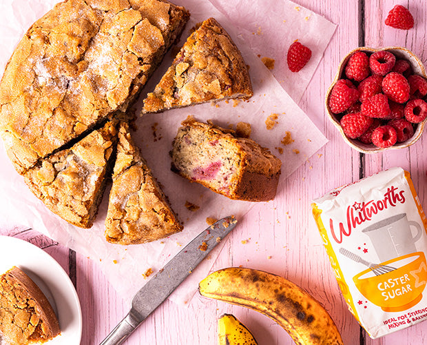 Banana and Raspberry cake, cut into slices with a bowl of fresh raspberries a banana and a pack of Whitworths Caster sugar.