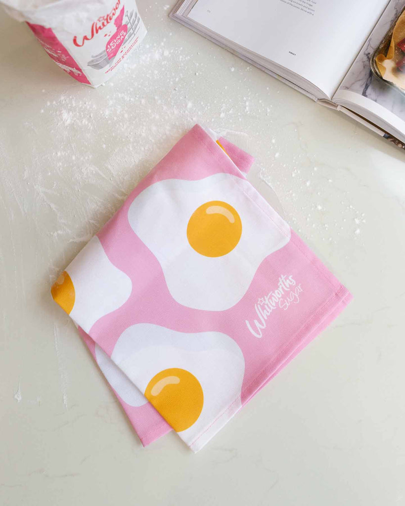 Pink Sunny Egg Tea towel folded on a table covered in icing sugar