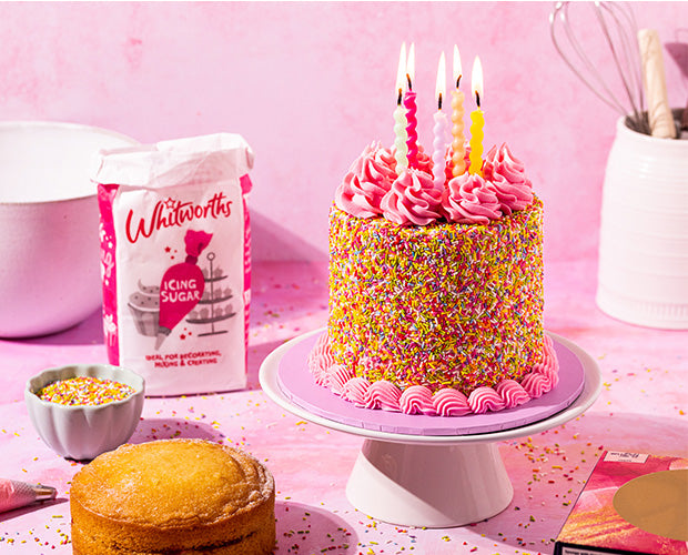 A Celebration Fake bake cake, smothered in Sprinkles, Pink piping around the base, Pink Swirls on the top, Lit candles, Whitworths Icing bag of sugar to the side along with a bowl of sprinkles and a baking bowl.