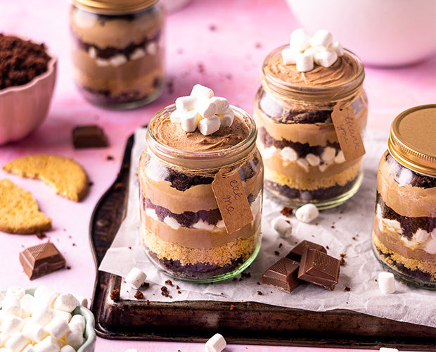 Glass jars filled with layers of Cake, Biscuits and Marshmallows, topped off with small marshmallows, surrounded by chunks of Chocolate and biscuits, bags of Whitworths golden Caster and Whitworths Dark soft Brown in the background.