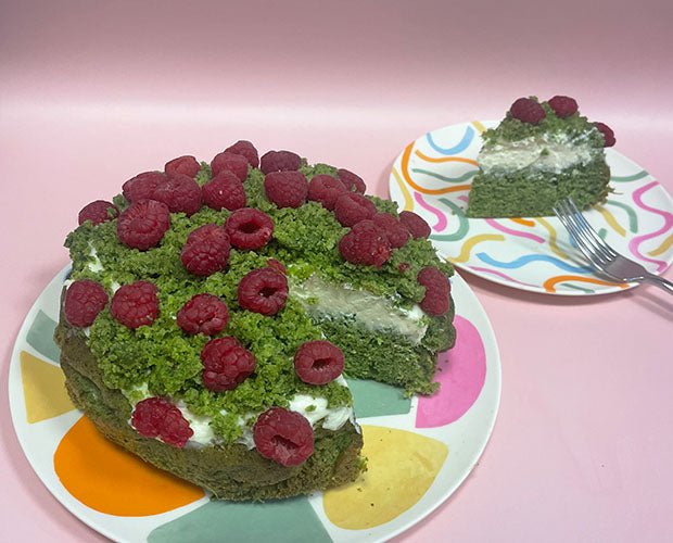 A layered green cake topped with crumbs to look like moss, filled with whipped cream and topped with fresh raspberries