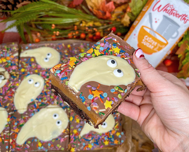 A festive Halloween dessert featuring layered Millionaire Brownie Bars with a fudgy brownie base, creamy caramel, and a chocolate topping decorated with playful chocolate ghosts. Perfect for Halloween celebrations!