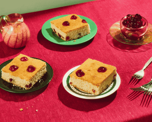 3 Slices of coconut cherry tray bake on colourful plates, next to a jug of fresh cream and bowl of glacier cherries